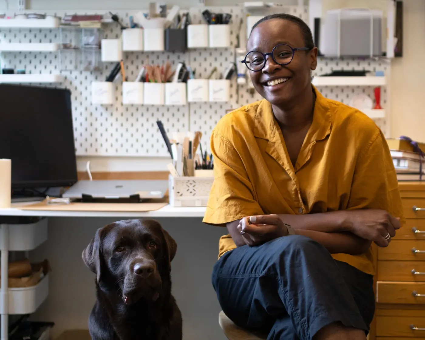 A woman with short hair and glasses sitting in an office, smiling, with a chocolate Labrador Retriever next to her. The office has white shelves with storage boxes and various supplies.