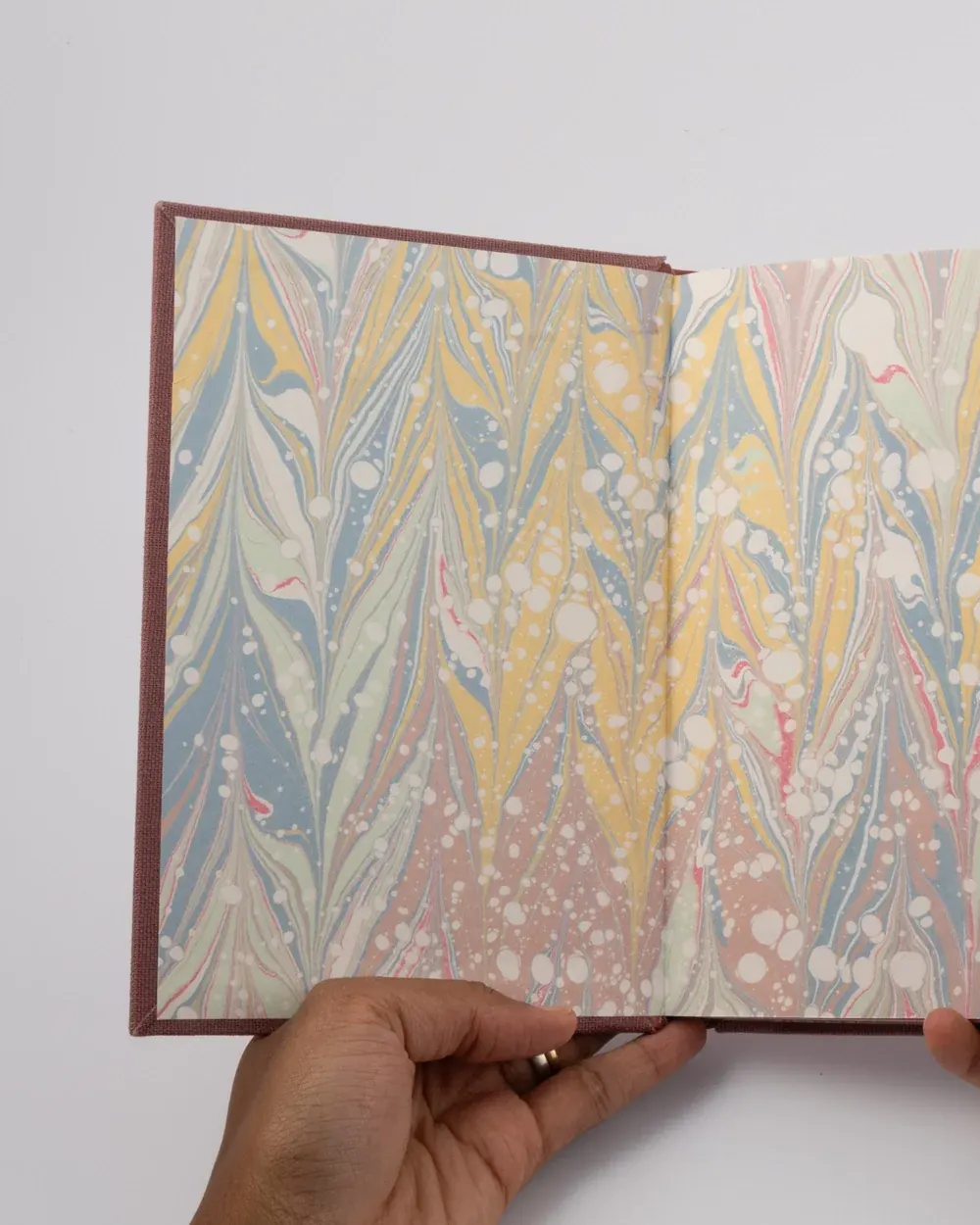 A hand holding an open book with marbled paper pages featuring colorful, swirling patterns in pastel shades of yellow, blue, green, pink, and white.