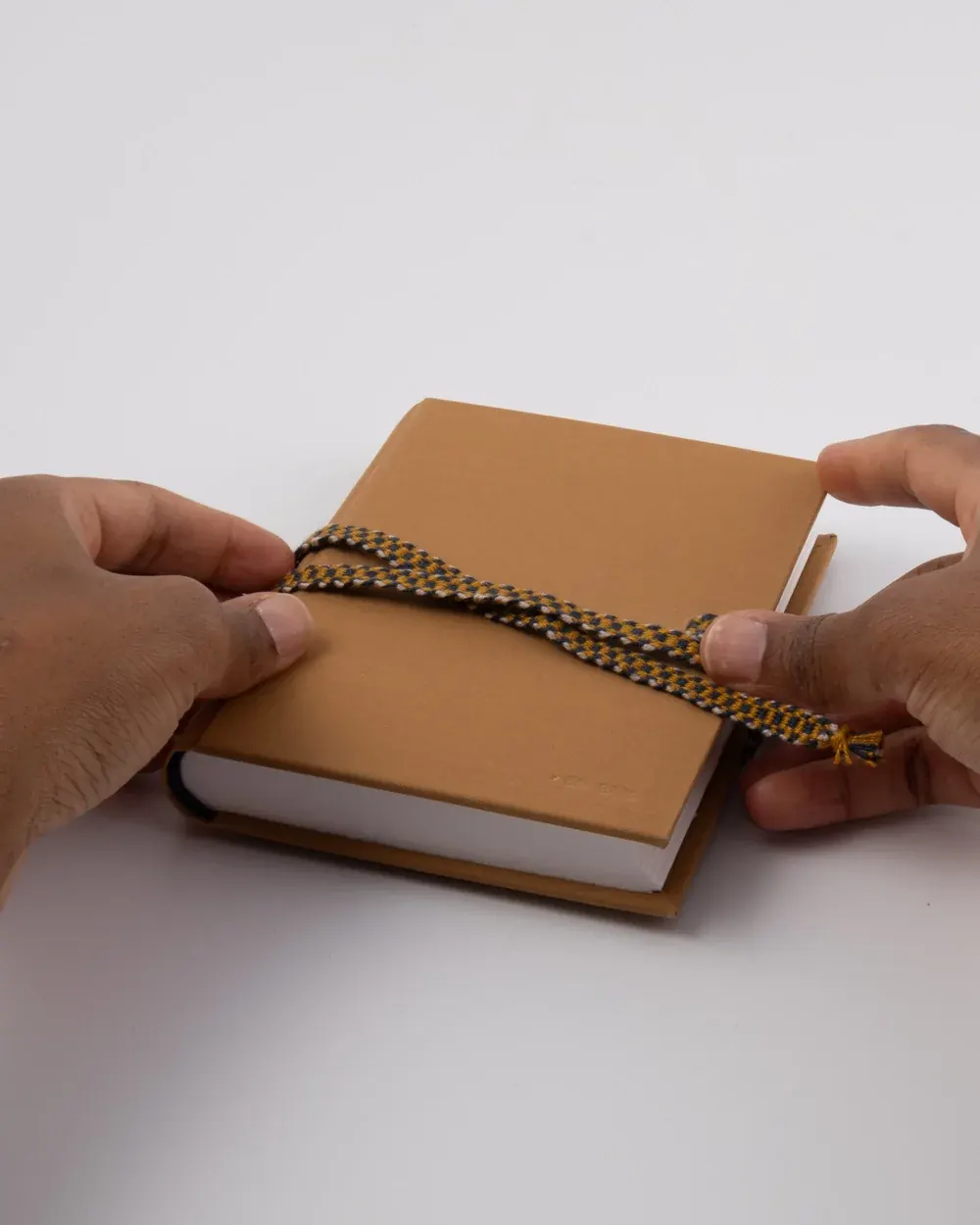 Hands holding a closed notebook with a brown cover, wrapped with a braided yellow and black cord, on a white surface.