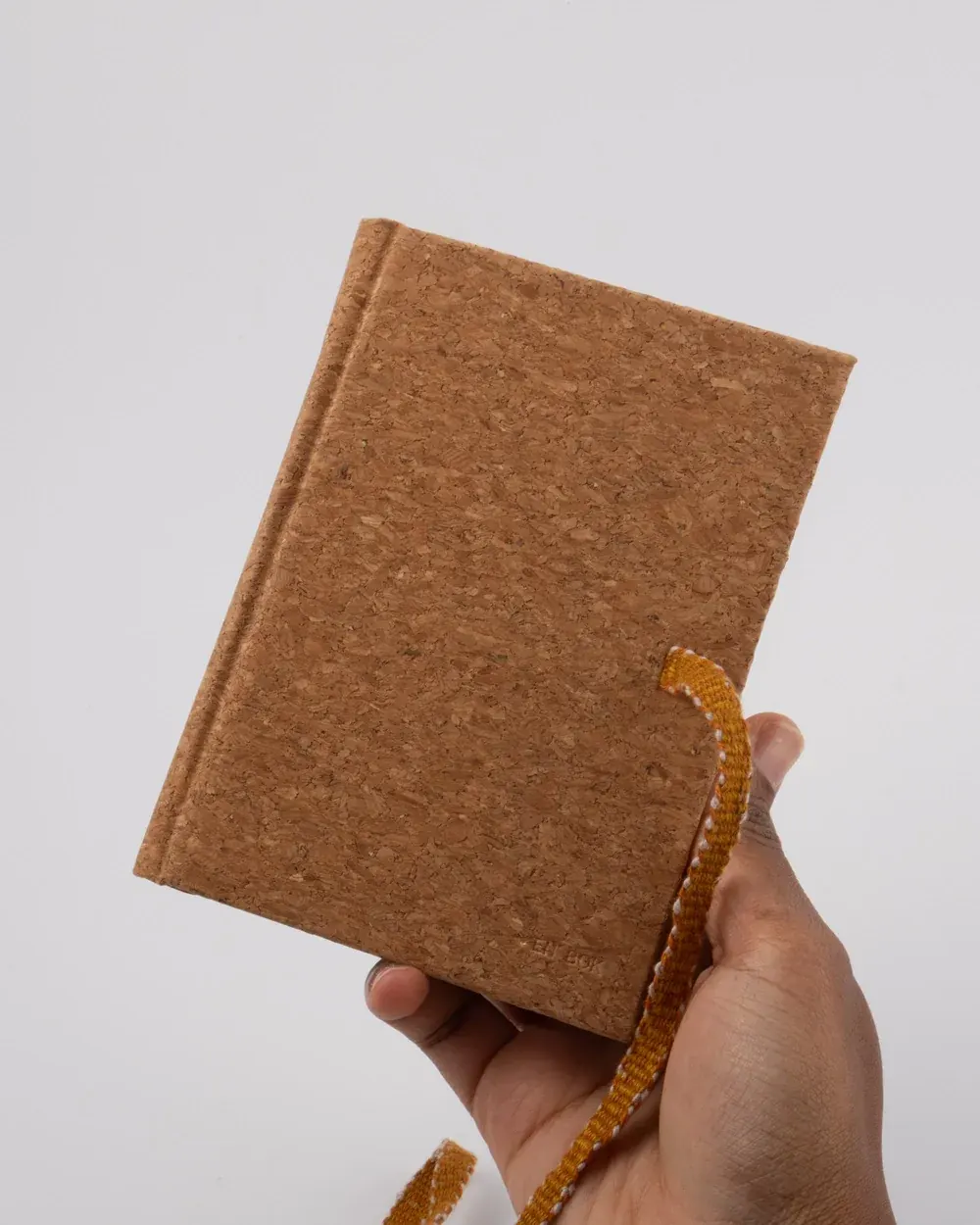 A hand holding a brown cork-covered notebook or journal with a yellow and white bookmark ribbon.
