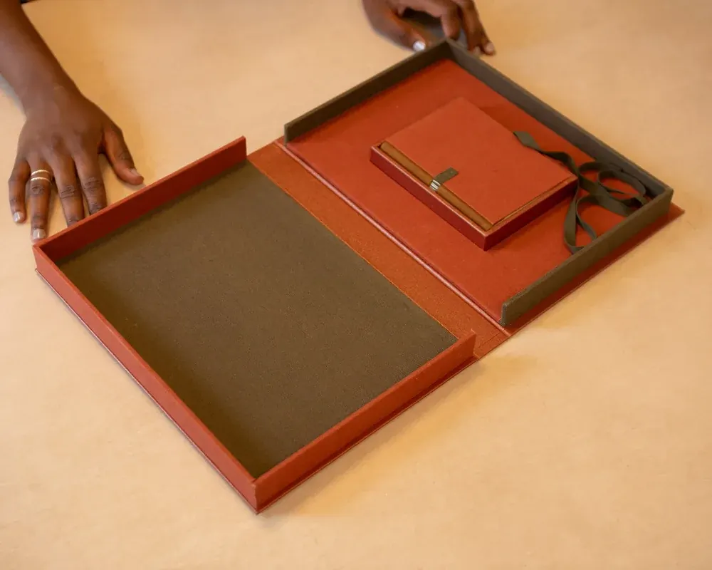 Open desk organizer with a red notepad, a brown notebook, and a black ribbon on a beige table.