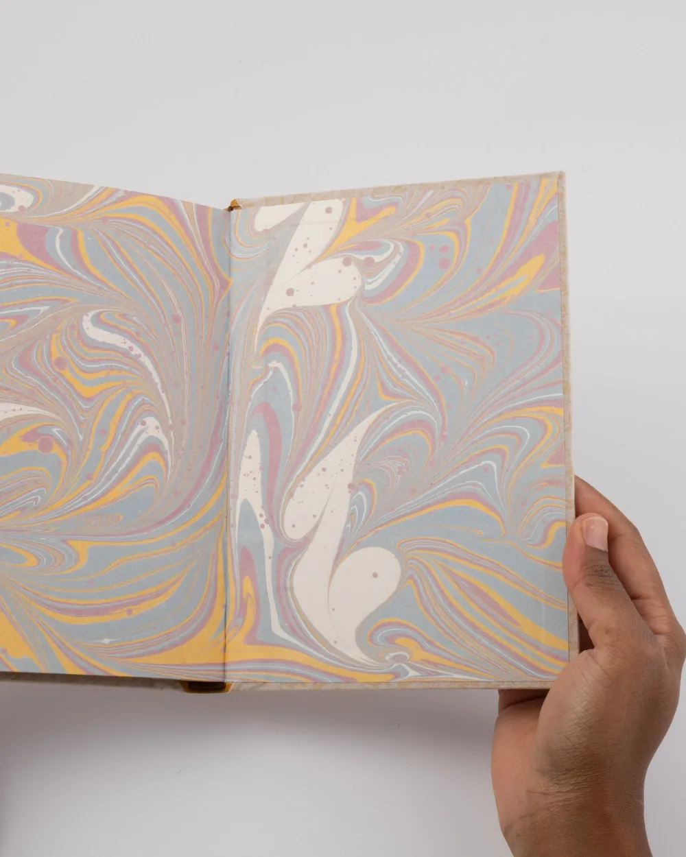 A person holding an open book with marbled paper inside, displaying swirling patterns of pastel colors including pink, yellow, blue, and white.