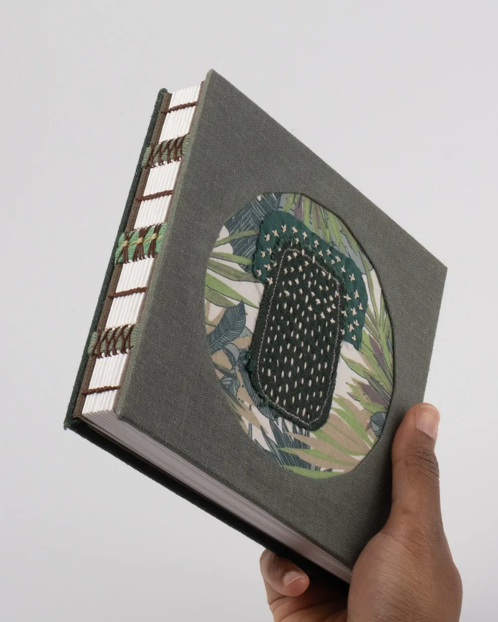 A hand holding a hardcover book with a fabric cover featuring a botanical design and an embroidered oval shape on the front.