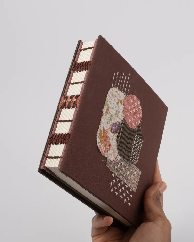 Hand holding a fabric-covered book with embroidered abstract art on the cover, white pages, and a textured spine.