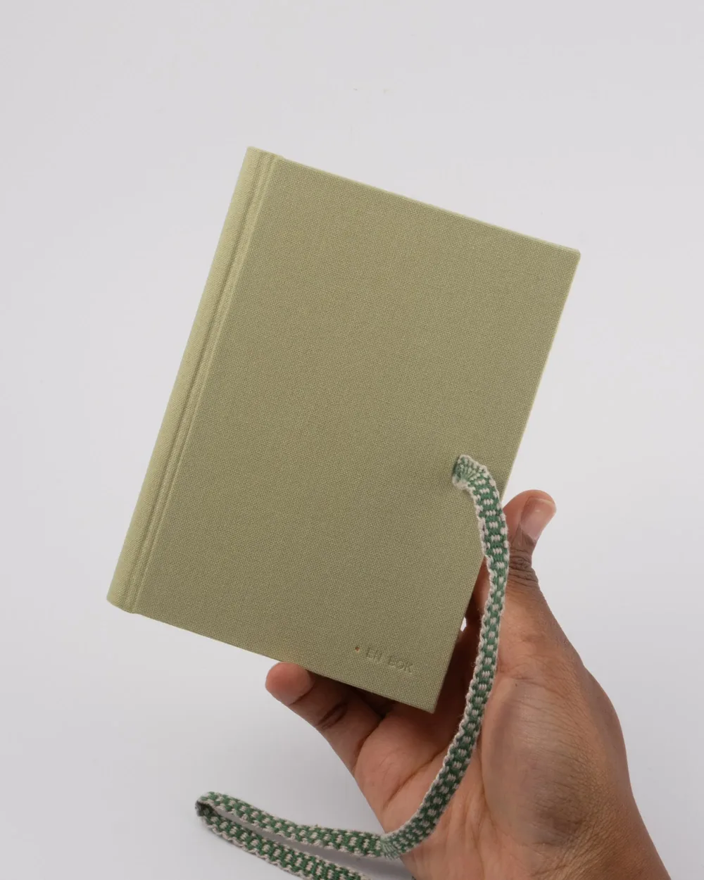 A person holding a closed, light green hardcover notebook with a green and white striped elastic band and a light green fabric cover against a plain white background.