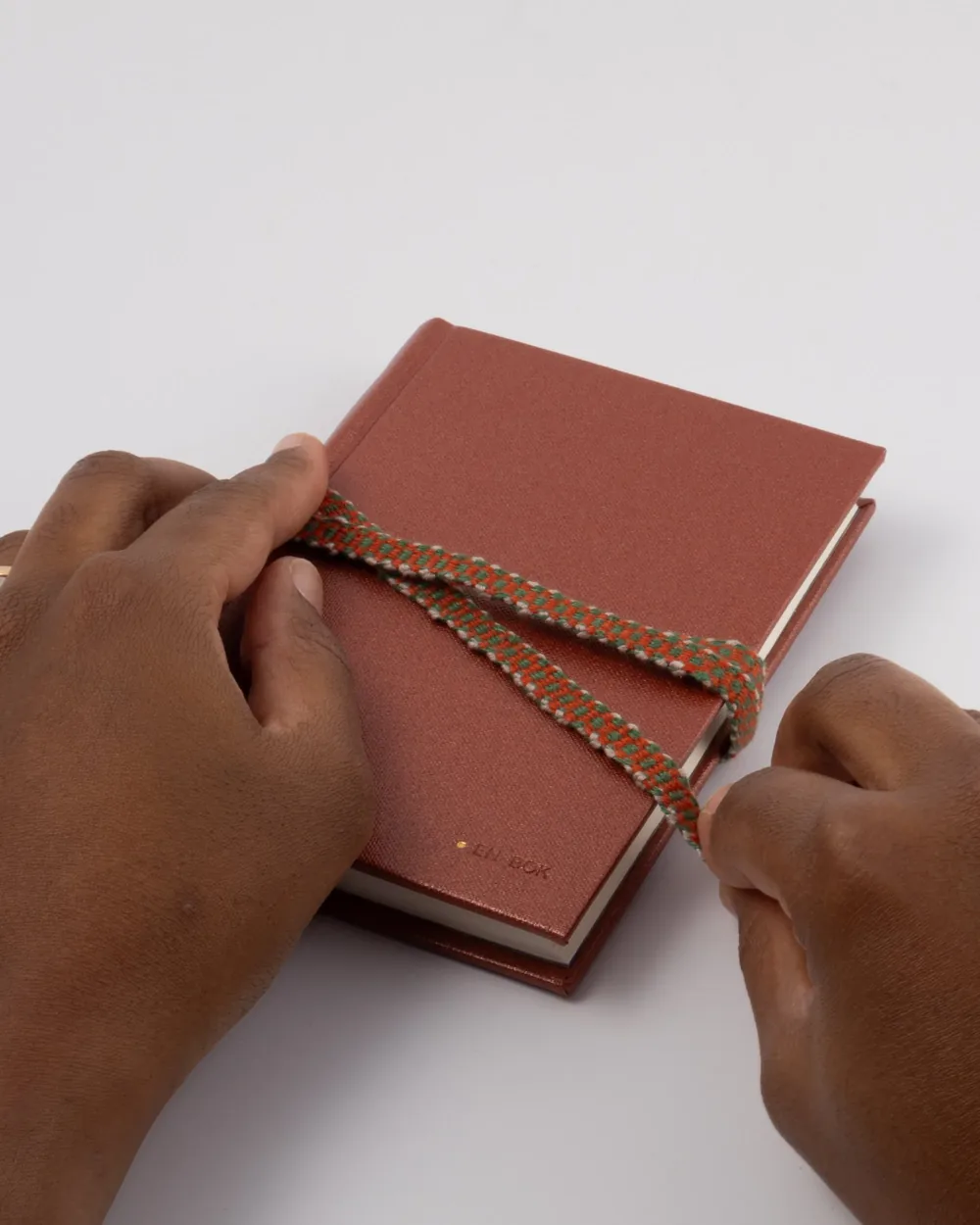 Hands wrapping a red hardcover book with a colorful braided bookmark on a white surface.