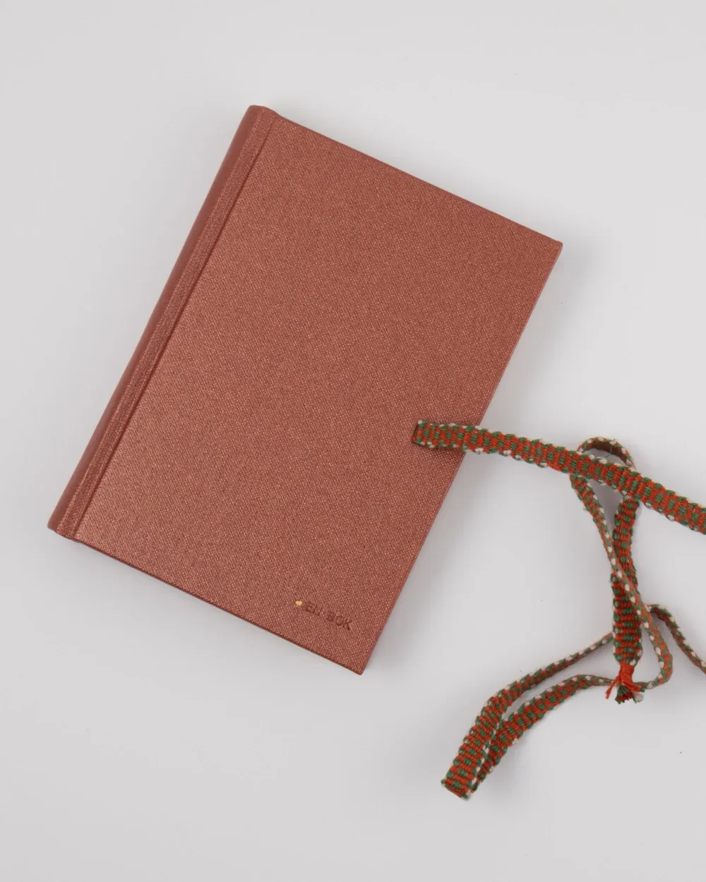 A closed pink hardcover book with a braided red and green ribbon bookmark on a white background.