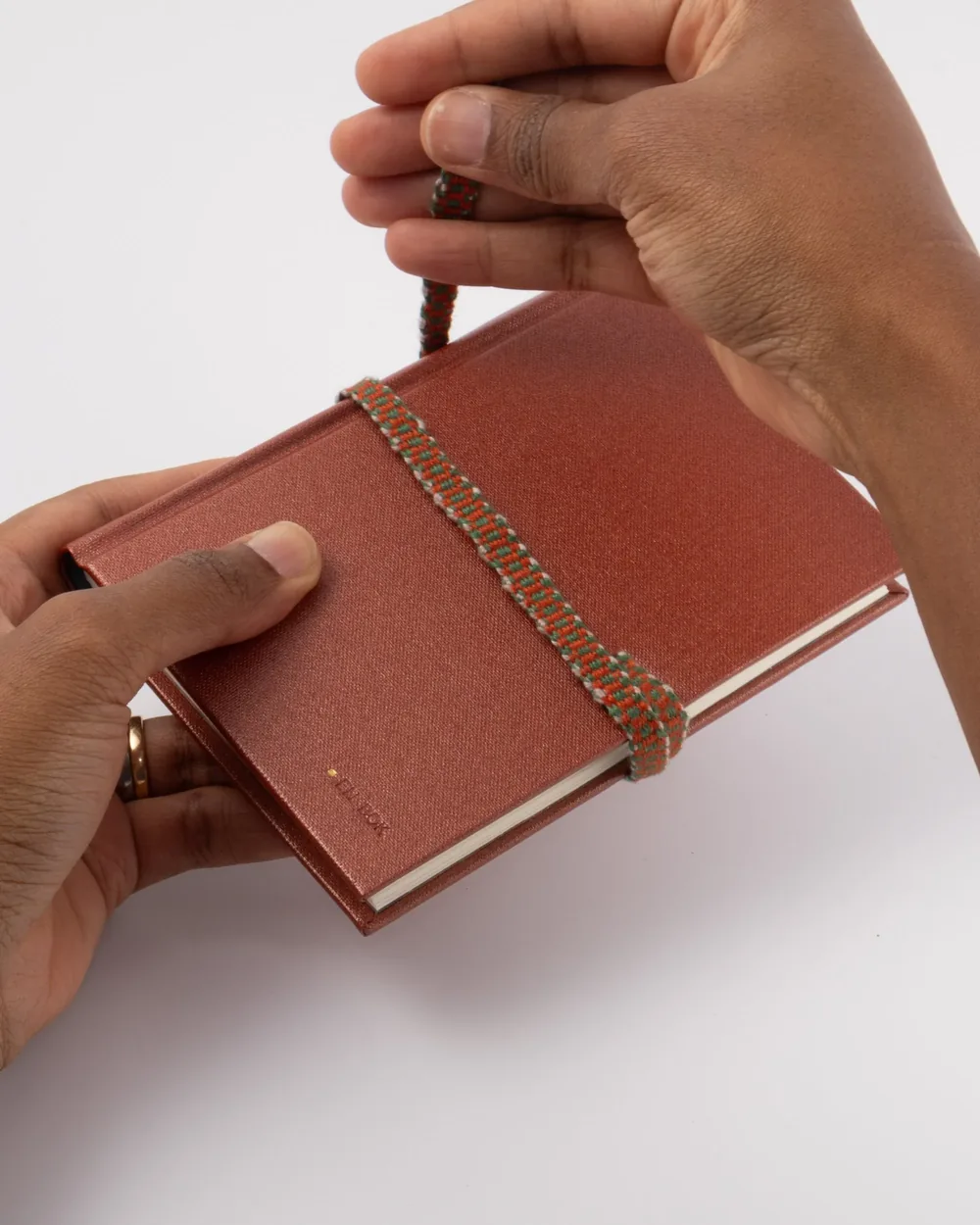 Hands holding a closed red journal with a colorful elastic band wrapped around it, on a white background.
