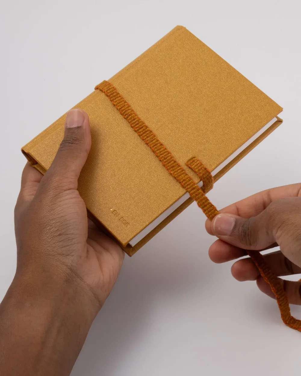 Hand holding a yellow hardcover notebook with an orange elastic strap on a white surface.