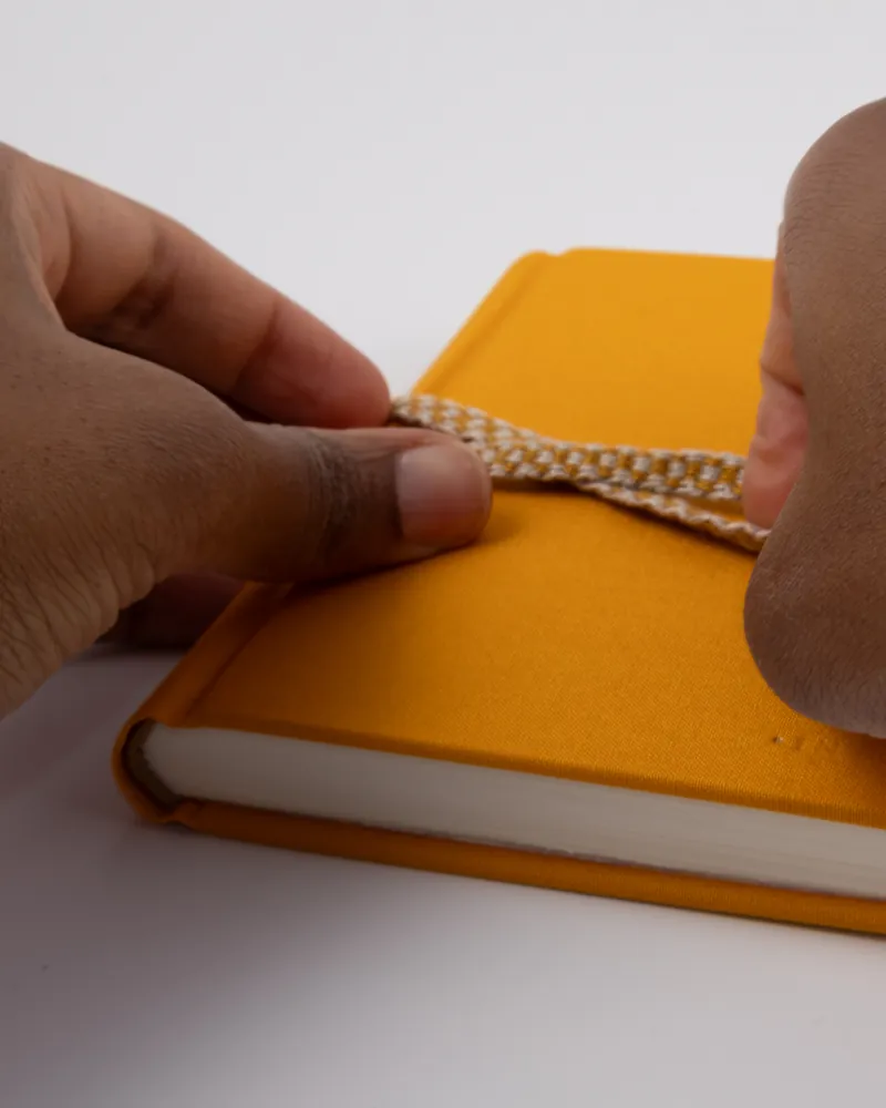 A pair of hands tying a yellow and white braided cord around an orange notebook with a yellow cover on a white surface.