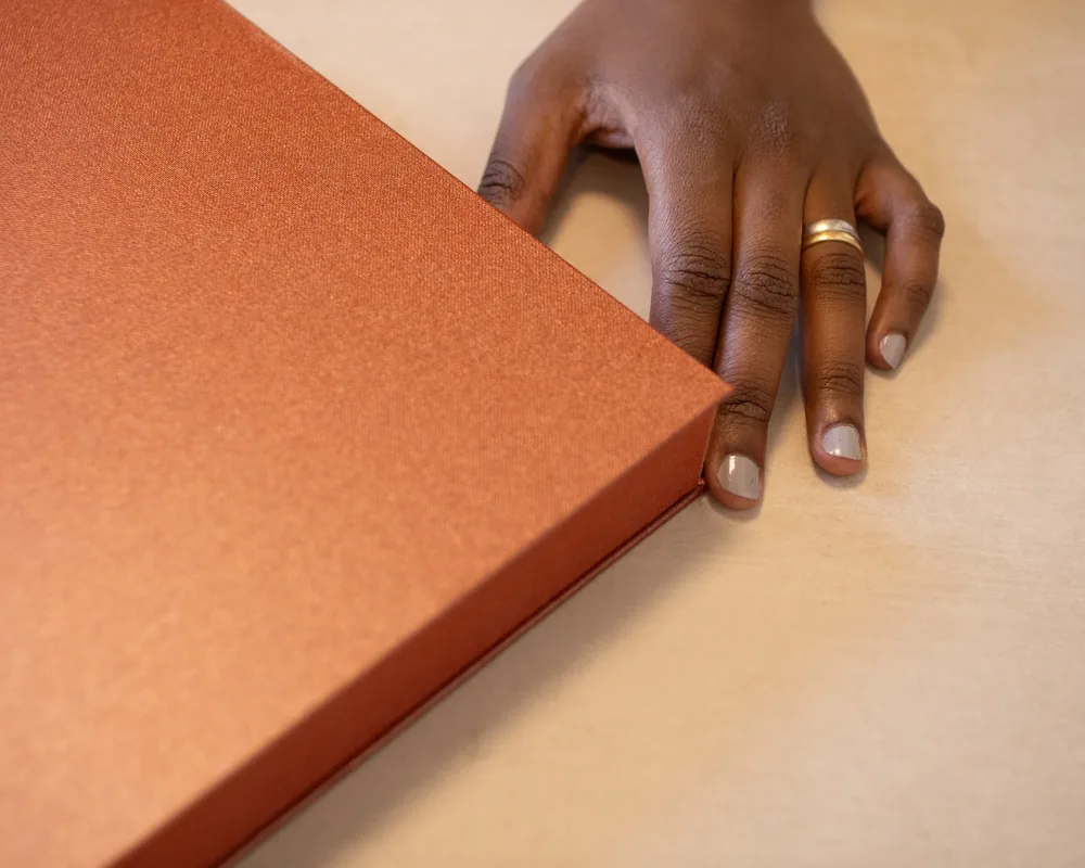 A person's hand with a wedding ring reaching out from behind an orange gift box on a beige surface.