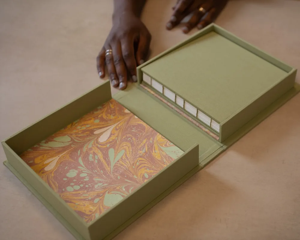 An open box with a green exterior and marbled interior, containing a pile of white books with gold edges. A person's hands are seen closing the box.
