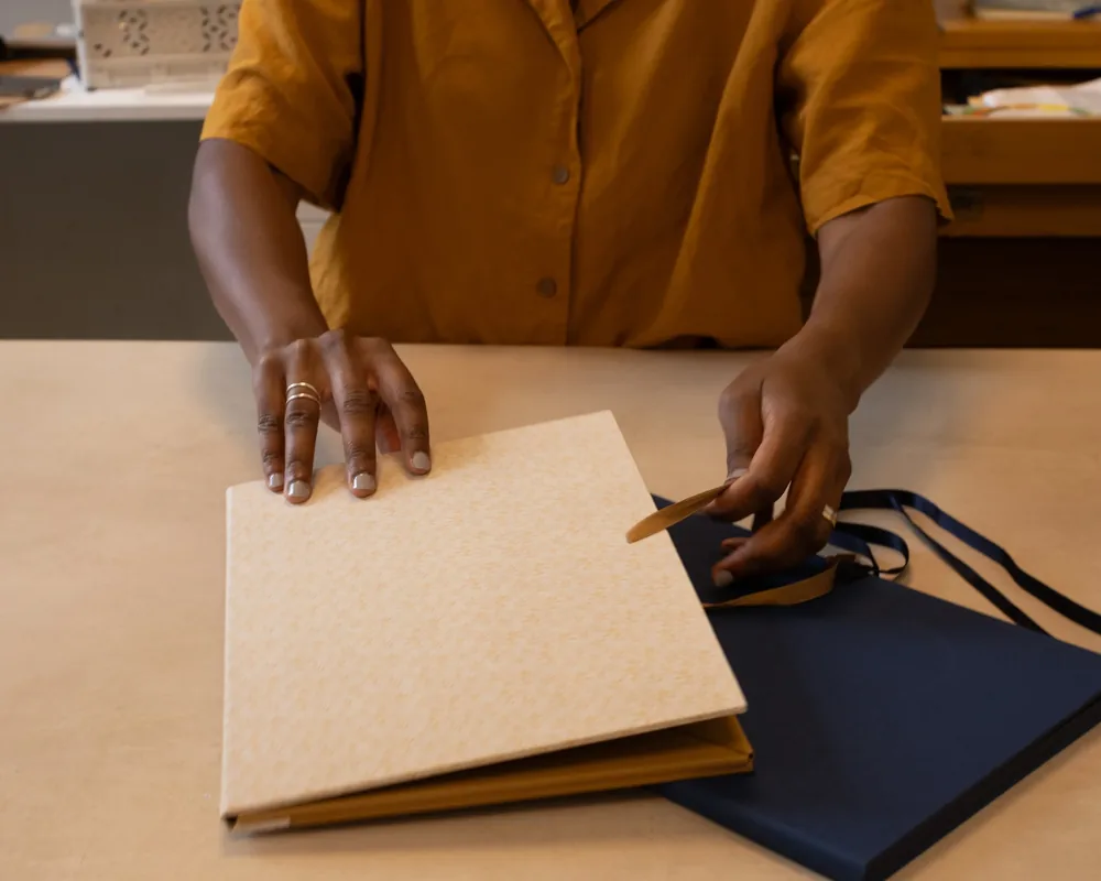 Person with dark skin wearing a mustard-colored shirt, holding a cardboard band, placing it on a closed book or notebook on a table.