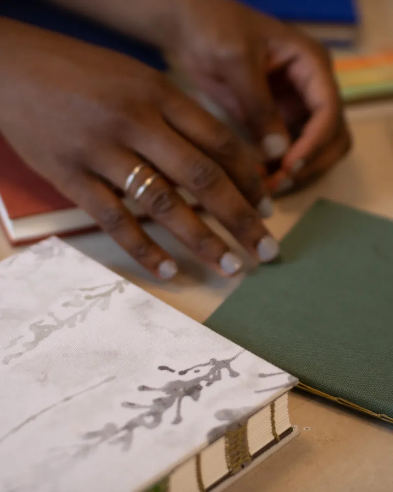 A person's hand with rings on fingers resting on a table among several notebooks and books.