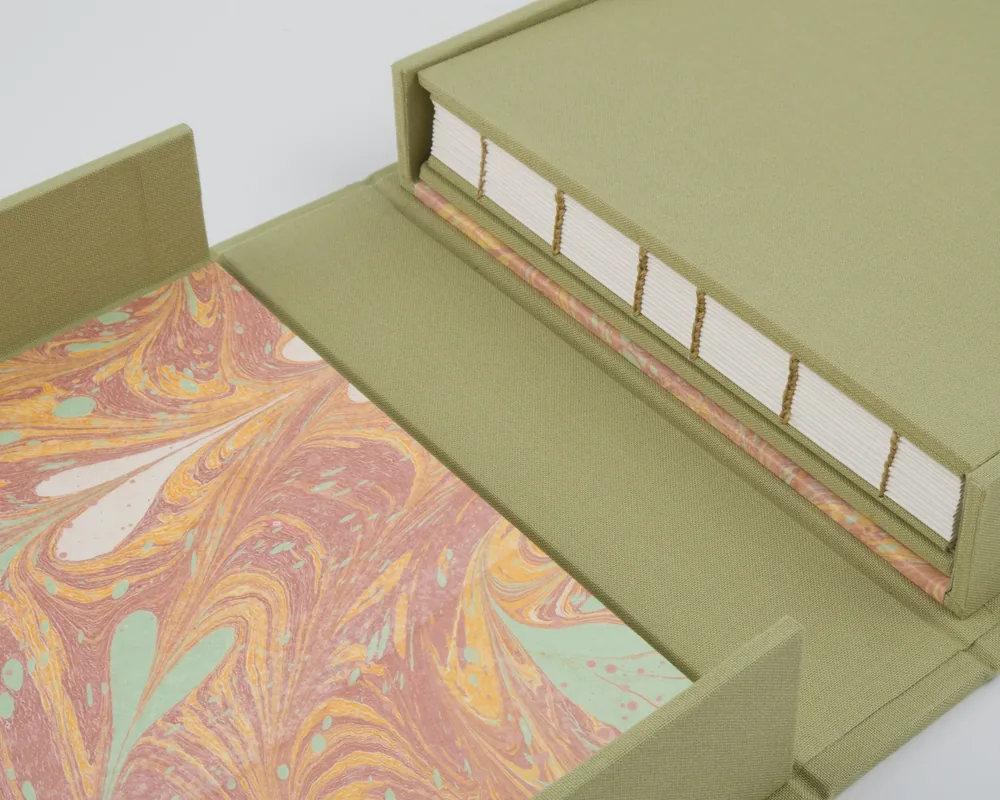 Open green fabric box with a marble-patterned interior and white books with gold ribbons inside.