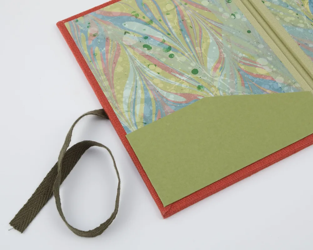 Close-up of a colorful, abstract-patterned fabric folder with a green paper pocket and a brown fabric strap on a white background.