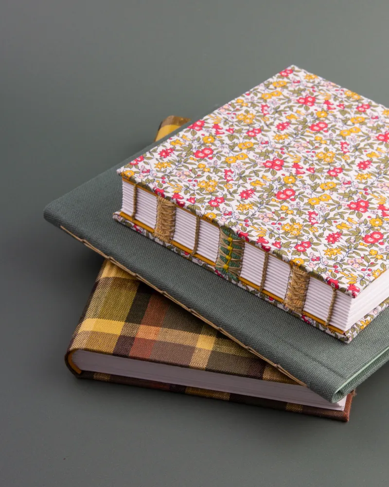 Three stacked fabric-covered notebooks with colorful floral and plaid patterns on a grey surface.