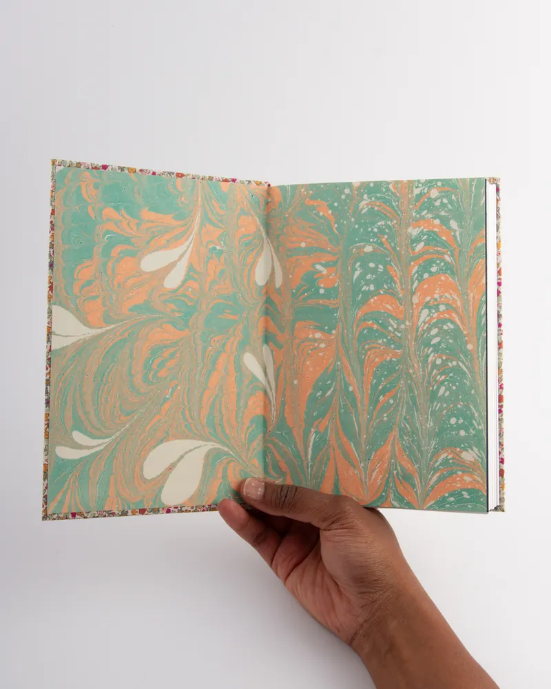 A person holding an open marbled paper book with colorful swirling patterns of green, orange, and white on a plain white background.