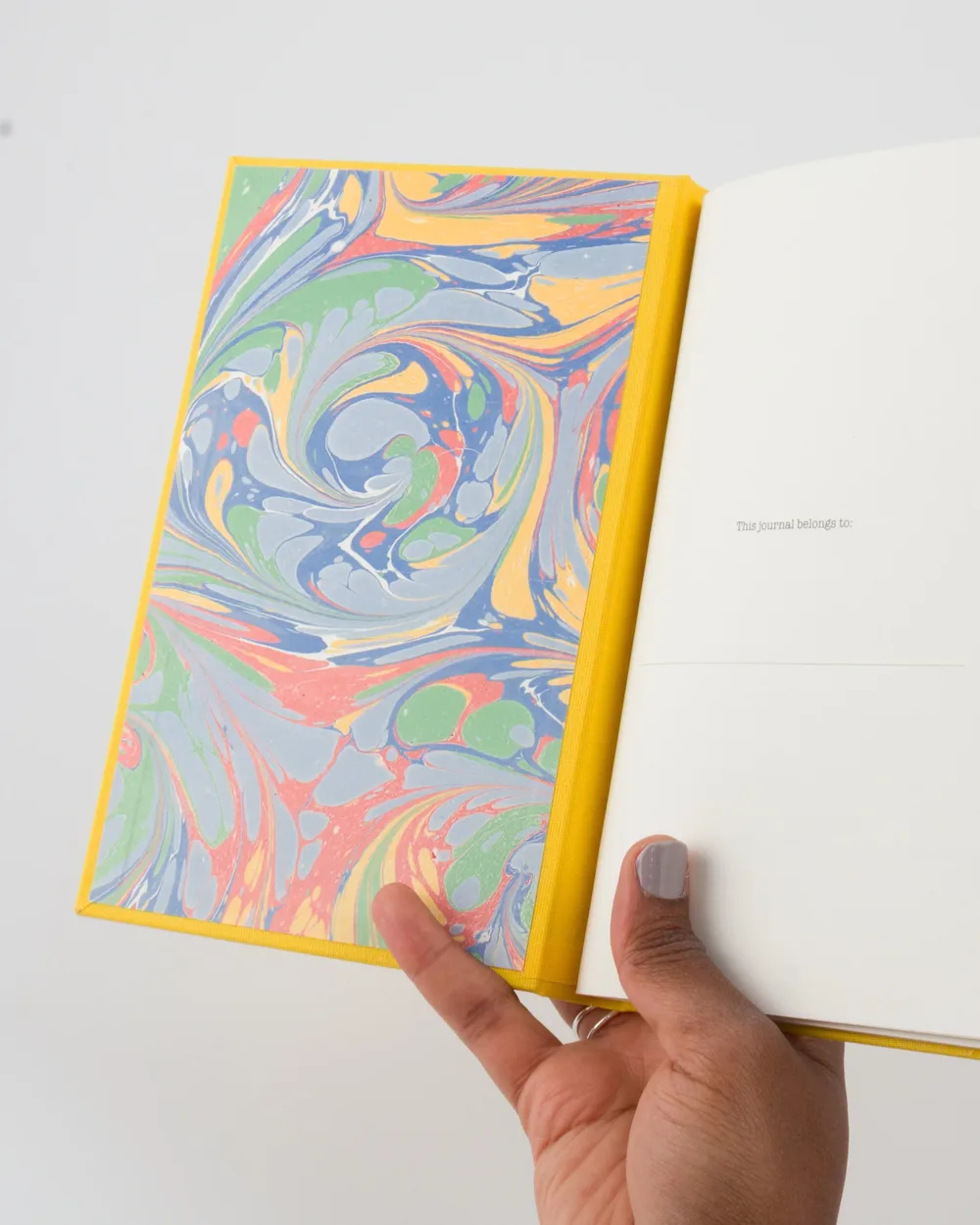 Open journal with marbled blue, green, pink, yellow, and orange splash pattern on the inside cover, held by a person with light gray nail polish.