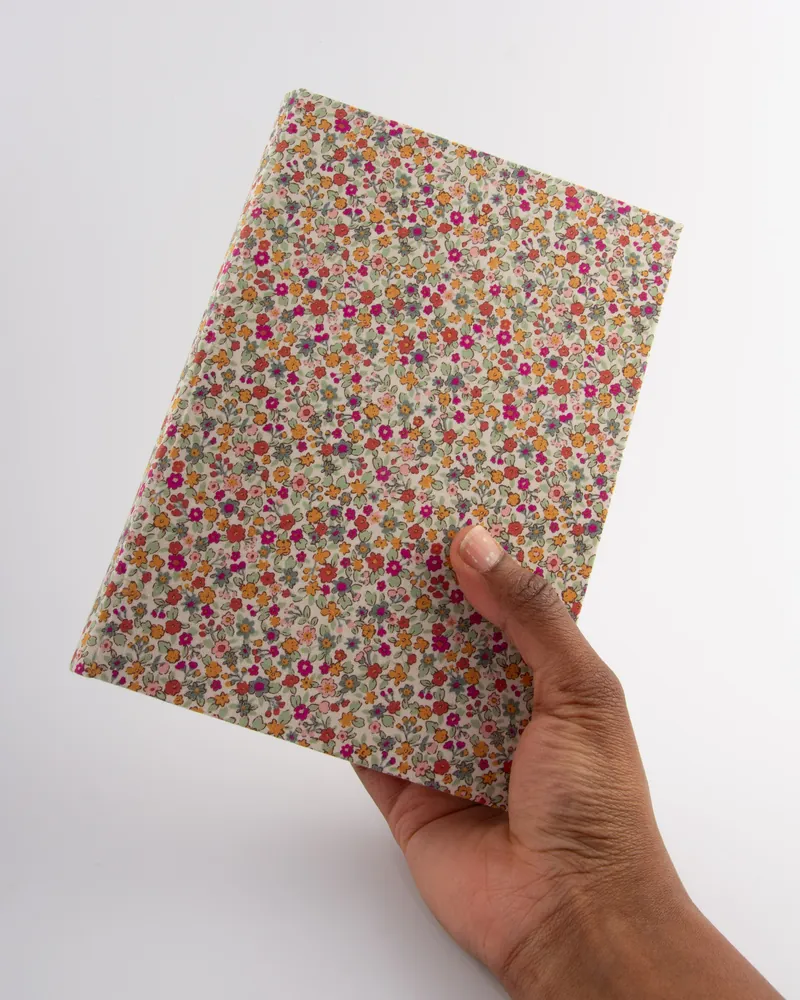 A hand holding a floral-patterned fabric folder or notebook with small pink, orange, purple, and green flowers on a white background.