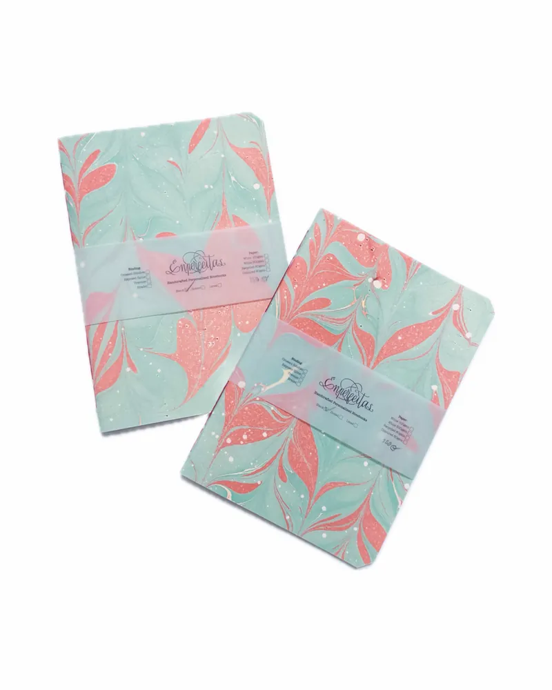 Two pastel-colored notebooks with marbled pattern covers in pink, mint green, and white, wrapped with transparent bands that feature the brand name 'Empreelitas'.