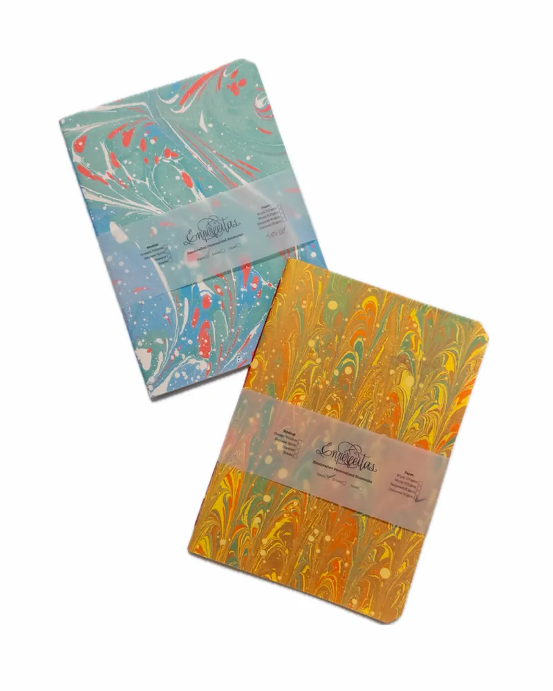 Two marbled handmade notebooks in colorful covers, one with blue and red swirls, and the other with yellow and orange swirls, each wrapped with a transparent band labeled 'Las Empreñetas'.
