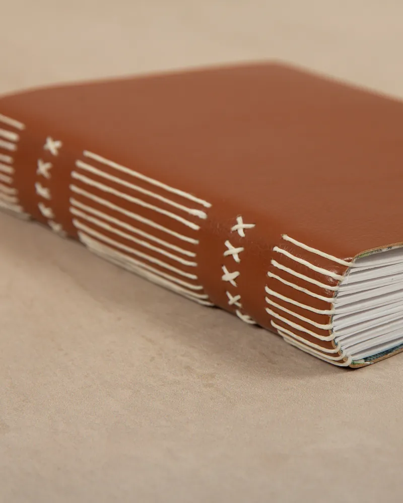 Close-up of a brown leather-bound book with white stitching on the spine, resting on a beige surface.