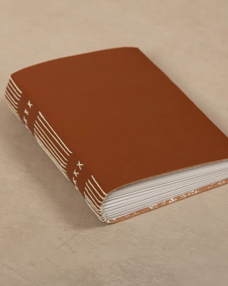 A brown notebook with white decorative lines and X's on the spine, lying on a beige surface.