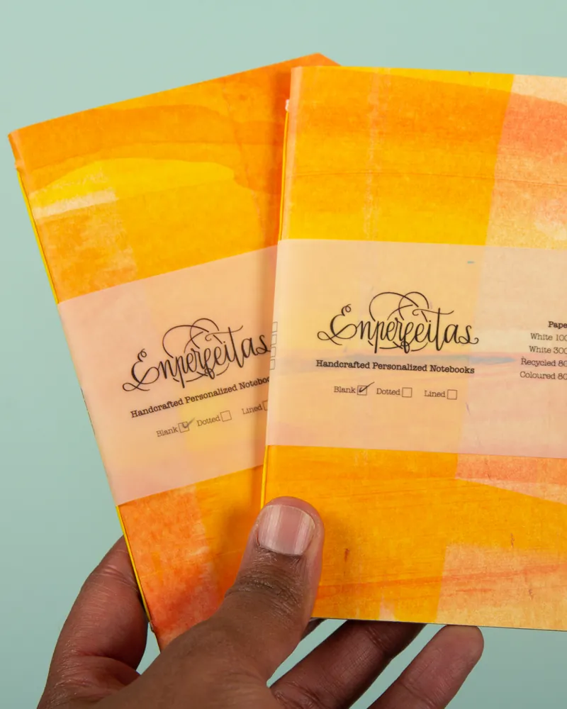 Two handcrafted personalized notebooks with orange-yellow abstract covers and white labels that read "Emperjetas." The labels include options for blank, dotted, and lined pages, with the blank option checked.