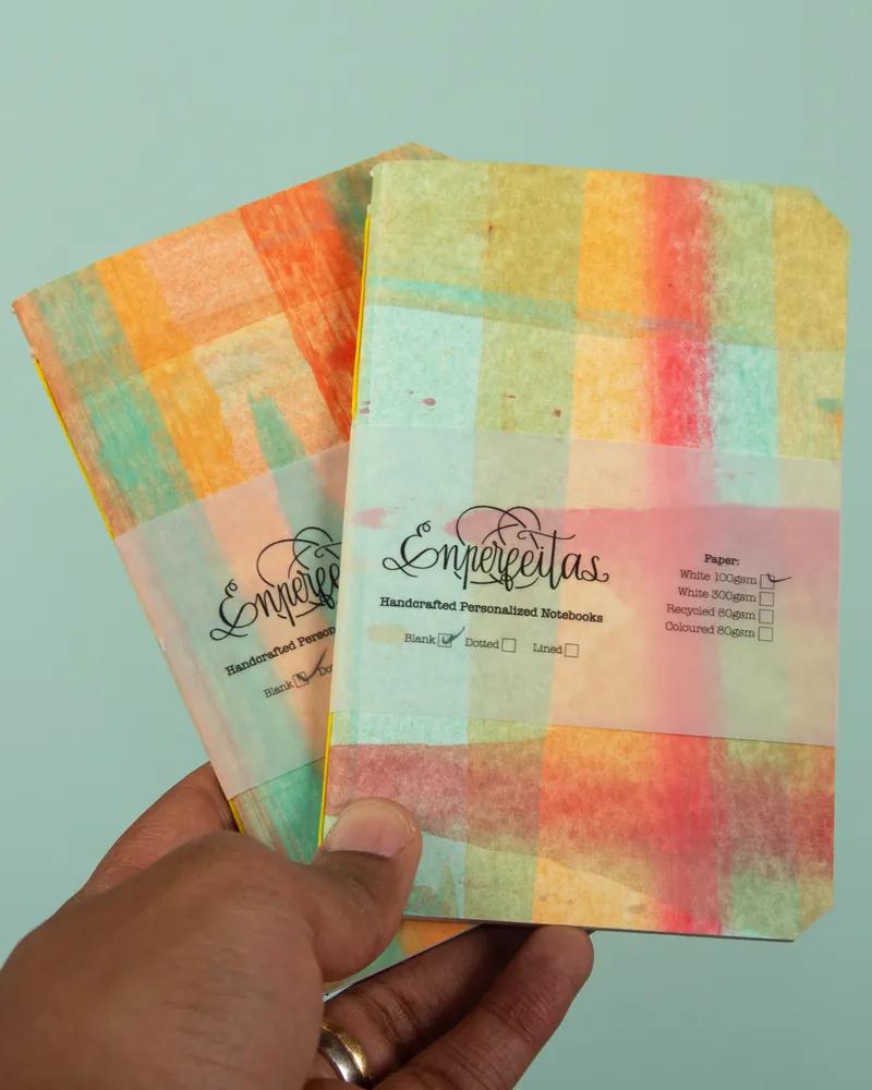 Two colorful, pastel-colored notebooks with a checkered pattern, held by a person with a ring on their finger, against a light green background.