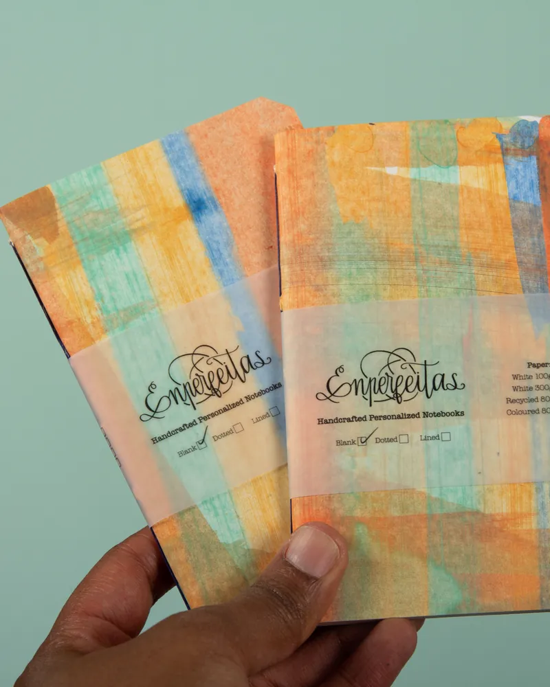 A hand holding two colorful, folded notebooks with a label that reads 'Enjepetlas Handcrafted Personalized Notebooks' and options for blank, dotted, or lined pages.