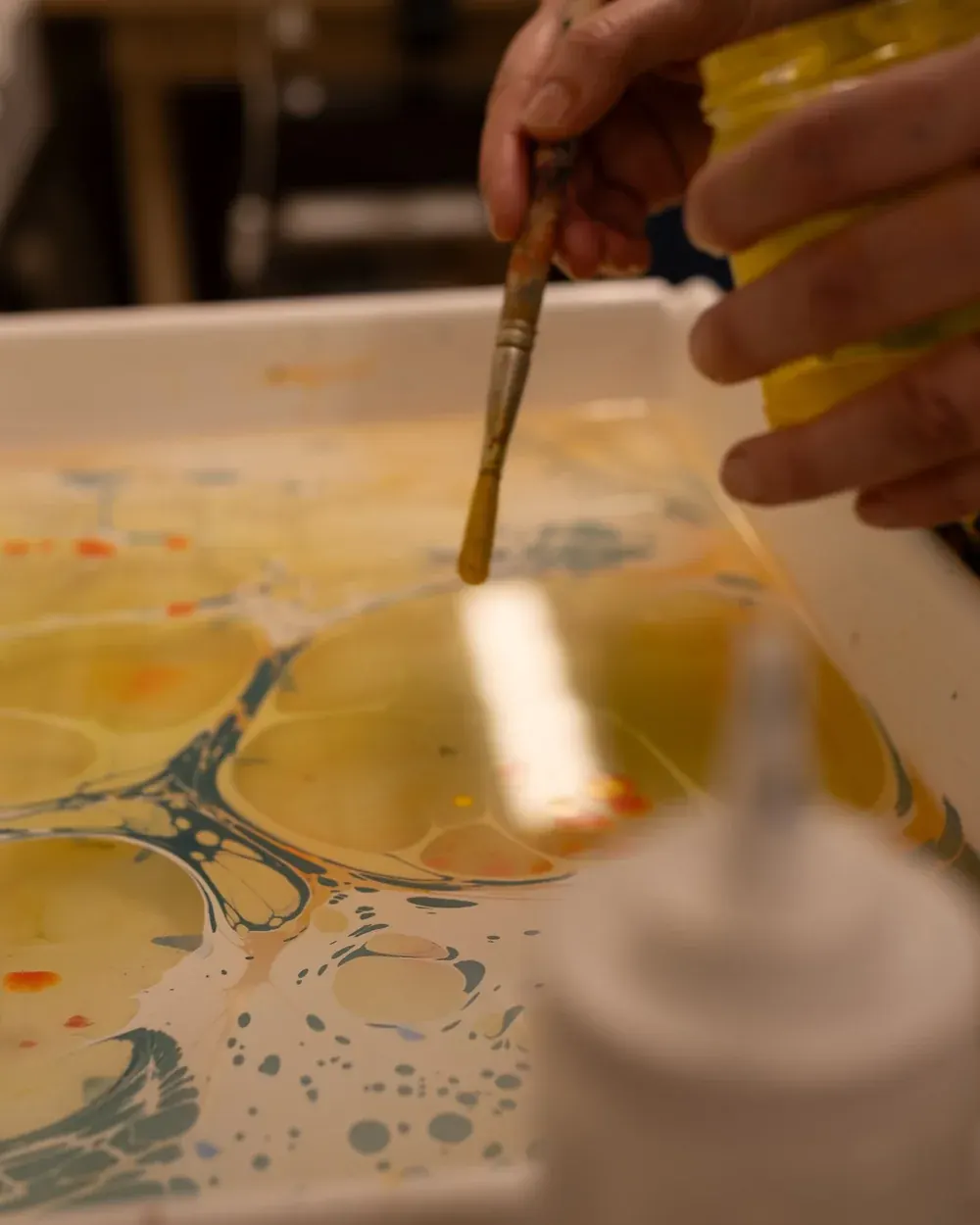 A hand marbling paper with orange, yellow, and blue inks in a shallow bath